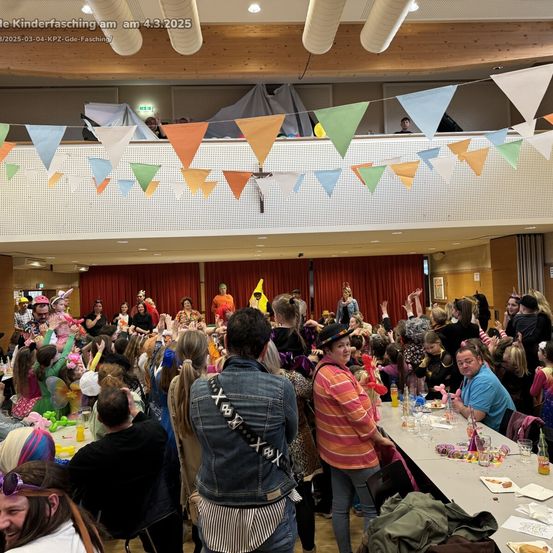 Eine große Gruppe von Menschen, einschließlich Kindern und Erwachsenen, versammelt sich in einem Raum für eine Kostümparty, viele tragen Kostüme und winken. Der Raum ist mit bunten Wimpelketten dekoriert und einem Tisch mit Essen und Getränken. Das Datum und die Uhrzeit sind oben links angezeigt.