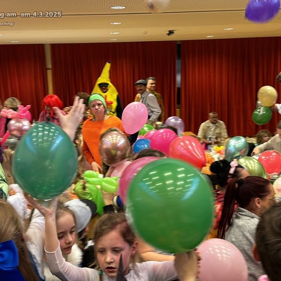Eine Menschenmenge in einem Festsaal, einige in Kostümen, halten Luftballons in verschiedenen Farben. Eine Frau vorne hält einen grünen Ballon, während andere um sie herum bunte Luftballons halten. Ein Mann in einer Bananen-Kostüm steht nahe einer roten Gardine.