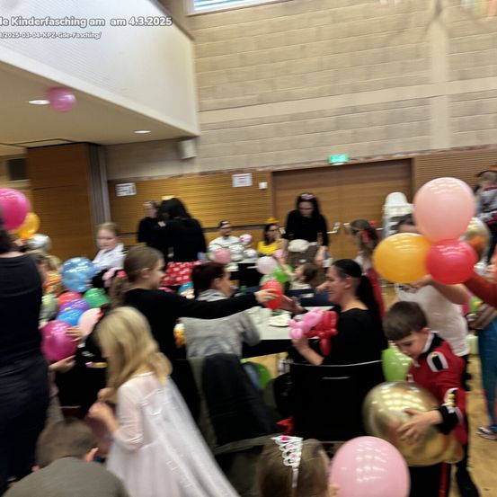 Eine Gruppe von Kindern und Erwachsenen versammelt sich in einem Saal und hält bunte Luftballons, um ein Ereignis zu feiern. Einige stehen, während andere um Tische herum sitzen. Die Wände sind aus Holz, und der Raum ist gut beleuchtet.