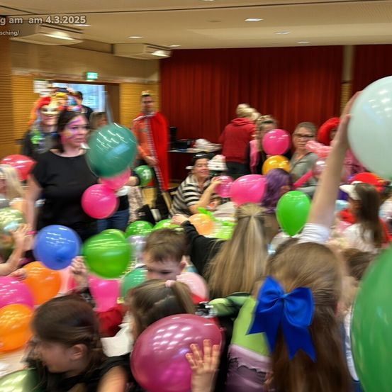 Ein lebhaftes Zimmer voller Kinder und Erwachsener, alle halten bunte Luftballons. Eine Frau in einem schwarzen Shirt mit Gesichtsbemalung steht vorne. Ein roter Vorhang und eine offene Tür sind im Hintergrund zu sehen.