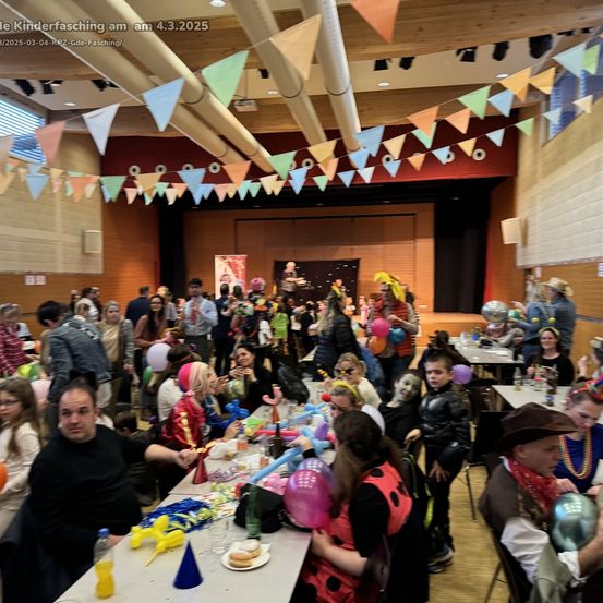 Eine große Versammlung beim Kinderfasching am 4. März 2025, gefüllt mit bunten Dekorationen, Luftballons und verschiedenen Kostümen. Die Menge sitzt an Tischen und genießt Essen und Getränke. Im Hintergrund ist eine Bühne mit einigen Personen zu sehen.