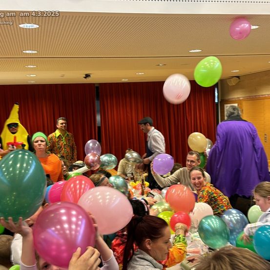 Eine Gruppe von Menschen feiert mit Luftballons, einige in Kostümen, in einem Raum mit roten Vorhängen. Eine Person in einem lila Kostüm steht hinten.