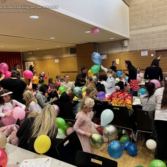 Eine große Gruppe von Menschen, wahrscheinlich bei einem Kinderfasching, versammelt sich in einem Raum. Sie halten und lassen bunte Luftballons steigen, einige in Kostümen. Der Raum ist mit Luftballons und Tischen dekoriert.
