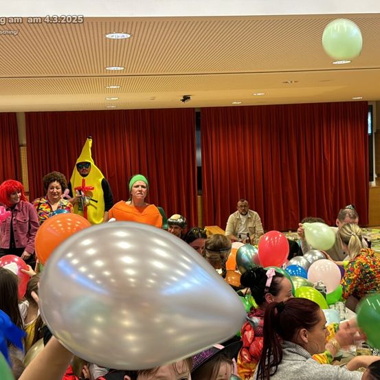 Eine Gruppe von Menschen in Kostümen, die Luftballons halten, versammelt sich in einem Raum mit roten Vorhängen und Deckenleuchten. Eine Person hält einen großen silbernen Ballon.