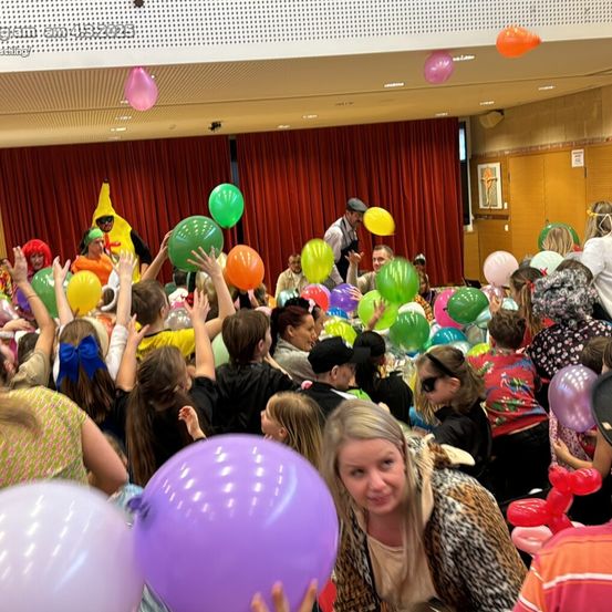 Eine große Gruppe von Menschen in einem Raum hält Luftballons hoch, einige von ihnen tragen Kostüme. Eine Person vorne hält einen lilafarbenen Ballon. Der Raum hat einen roten Vorhang und eine Holztür.