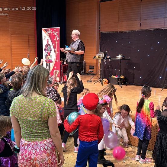Ein Zauberer führt auf einer Bühne vor einer Menge Kinder einen Zaubertrick vor. Der Zauberer hält eine Schüssel und scheint einen Zaubertrick zu machen. Die Kinder schauen zu und einige halten Luftballons. Die Bühne hat einen schwarzen Hintergrund mit Sternen.