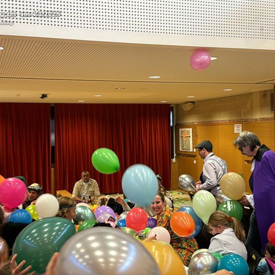 Eine Gruppe von Menschen feiert mit Luftballons in verschiedenen Farben. Sie stehen und sitzen in einem Raum mit roten Vorhängen, und ein Mann hält eine Menge Luftballons.