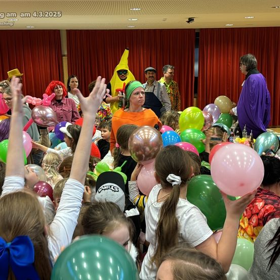 Eine Gruppe von Menschen bei einer Veranstaltung mit Luftballons und Kostümen, einige mit roten Perücken und Masken, eine Frau mit einer Bananen-Kappe und andere in bunten Outfits.