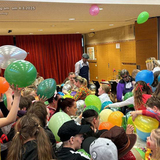 Eine große Gruppe von Menschen in einem Raum, einige in Kostümen, halten und werfen Luftballons. Ein Mann steht in der Nähe einer Tür.