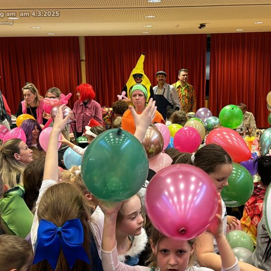 Eine Gruppe von Kindern in einem Raum mit vielen Luftballons, einige in Kostümen. Eine Frau mit einer Bananenmütze und Sonnenbrille steht hinter ihnen. Es gibt Erwachsene im Hintergrund, und rote Vorhänge sind im Hintergrund.