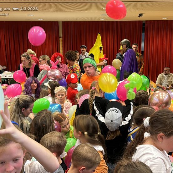 Eine Gruppe von Menschen, einschließlich Kindern, versammelt sich in einem mit Ballons gefüllten Raum. Einige Erwachsene tragen Kostüme, und der Raum hat rote Vorhänge und einen Tisch mit Gegenständen. Das Datum und die Uhrzeit sind 4.3.2025 um 4 Uhr morgens.