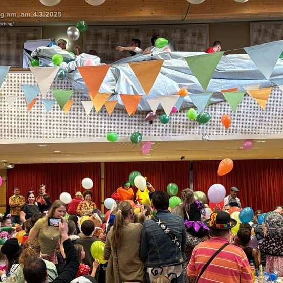 Eine lebhafte Indoor-Veranstaltung zeigt eine Menschenmenge in Kostümen, die Ballons halten. Eine Person auf einer Plattform mit Luftballons blickt auf die Menge. Bunte Banner und Flaggen hängen von der Decke.