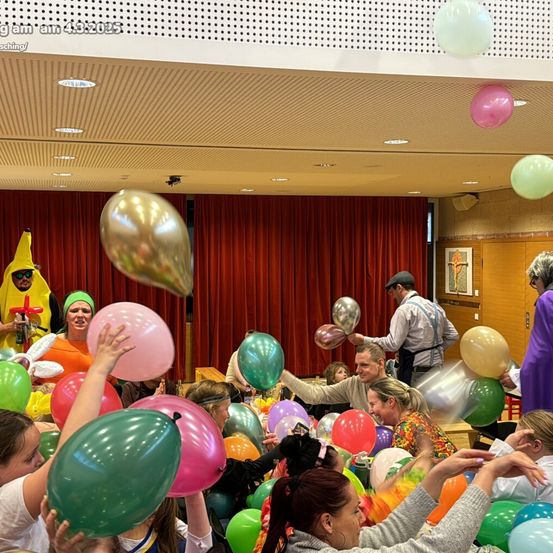 Indoor-Veranstaltung mit vielen Luftballons; Menschen in Kostümen, einschließlich eines Mannes in einer Bananen-Verkleidung, lachen und halten Luftballons. Rote Vorhänge im Hintergrund.