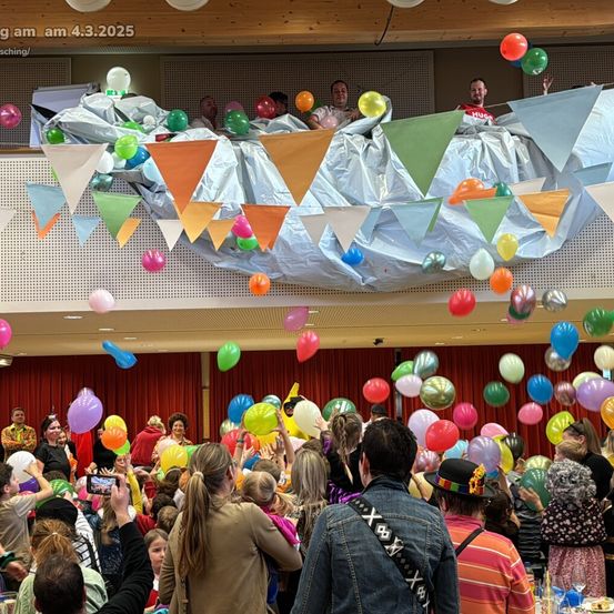 Eine Gruppe von Menschen in einem großen Saal feiert mit einer festlichen Atmosphäre. Es gibt viele bunte Ballons, die in der Luft schweben. Oben auf dem Balkon halten einige Menschen Ballons und scheinen an der Feier teilzunehmen. Die Menge unten ist vielfältig, mit verschiedenen Personen in unterschiedlichen Kostümen, die Ballons hochhalten.