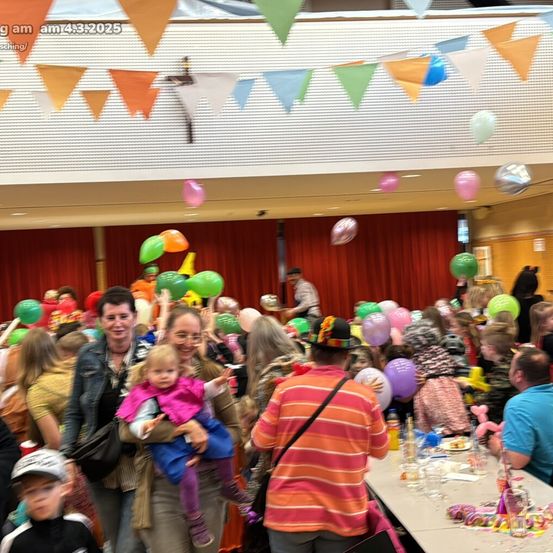 Ein Raum voller Menschen, einige halten Luftballons und feiern ein Ereignis. Eine Frau hält ein junges Mädchen, beide lächeln. Ein Mann in einem gestreiften Shirt steht in der Nähe. Tische mit verschiedenen Gegenständen, wie Gläsern und Essen, sind vorhanden.