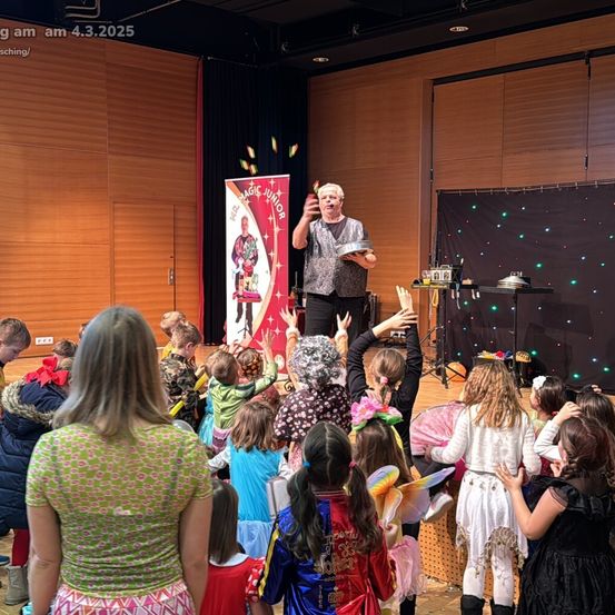 Ein Zauberer führt auf einer Bühne vor einer Menge von Kindern in Kostümen auf, die zusehen. Die Bühne hat eine Holzwand und einen schwarzen Hintergrund mit Lichtern.