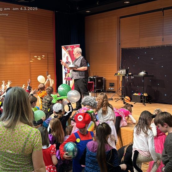 Ein Zauberer führt auf einer Bühne vor einer Menge von Kindern in Kostümen vor. Der Mann hält eine Schüssel und einen Zauberstab. Die Kinder halten Luftballons. Die Bühne hat einen Hintergrund mit Sternen und ein Banner mit dem Poster des Zauberers.