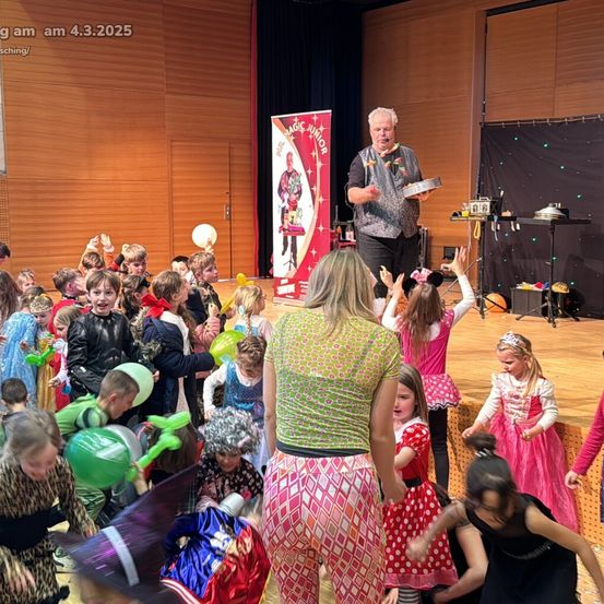 Ein Zauberer führt eine Vorstellung auf der Bühne mit Kindern in bunten Kostümen und Luftballons durch, mit einem Banner und einem Lautsprecher im Hintergrund.