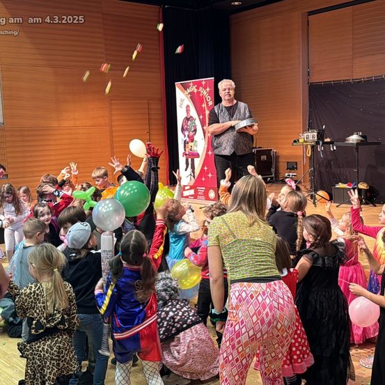 Ein Zauberer führt eine Ballontrickshow für eine Gruppe von Kindern auf der Bühne vor, mit Holzwänden und schwarzem Hintergrund.