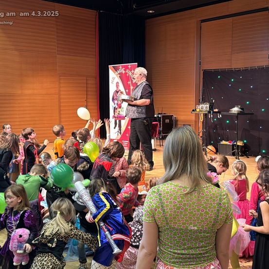 Ein Zauberer führt auf der Bühne vor einer Menge von Kindern in Kostümen auf. Er hält eine silberne Schüssel und eine Karte, während die Kinder ihre Hände heben und Luftballons halten.