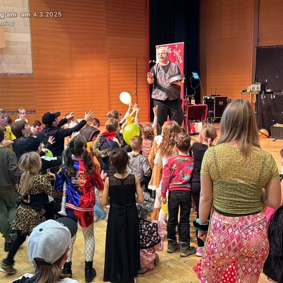 Ein Mann führt auf der Bühne vor einer Menge Kinder in Kostümen auf. Der Mann hält einen Hut und eine Kugel. Die Kinder jubeln und heben ihre Hände. Die Bühne ist mit einem Banner und einem Vorhang dekoriert. Es gibt Tische und Ausrüstung an der Seite. Das Bild hat einen Wasserzeichen.
