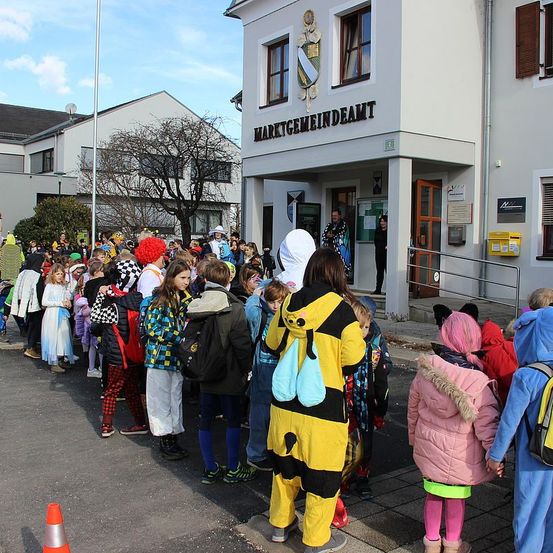 Eine Gruppe von Kindern in Kostümen steht in einer Reihe vor einem Gebäude mit dem Schild 'MARKTGEMEINDEAMT' oben. Einige tragen Rucksäcke.