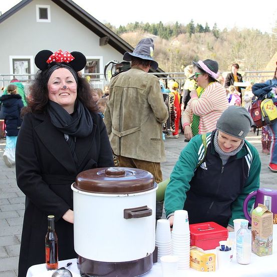 Eine Frau mit Minnie-Maus-Haarband steht neben einem Crockpot bei einer Outdoor-Veranstaltung. Neben ihr sitzt eine weitere Frau, lächelt und hält eine rote Box. Im Hintergrund sind ein Mann mit Hexenhut und andere in festlicher Kleidung zu sehen.