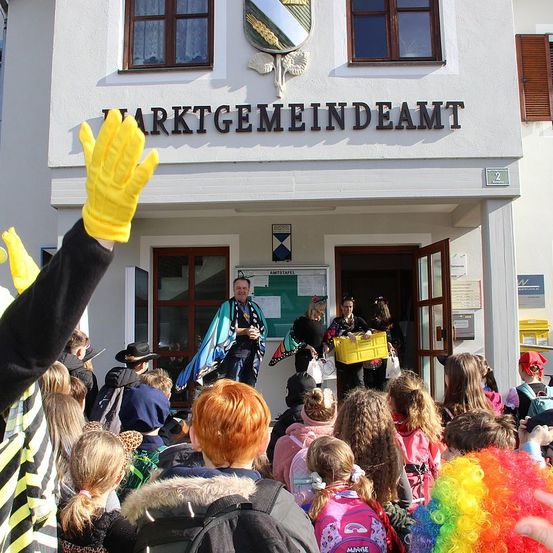 Eine Gruppe von Kindern in Kostümen und Rucksäcken versammelt sich vor einem Gebäude mit einem Schild, das 'Marktgemeindeamt' steht. Das Gebäude hat zwei Türen, von denen eine offen ist, und ein Fenster mit einem weißen Vorhang. Eine Person in einem gelben Handschuh hebt die Hand.