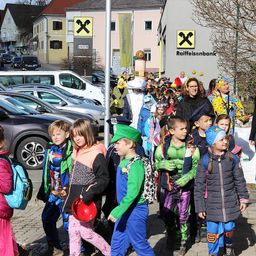 Eine Gruppe von Kindern in Kostümen geht mit geparkten Autos und einem Schild für Raiffeisenbank die Straße entlang. Sie werden von Erwachsenen begleitet und sind wahrscheinlich auf dem Weg zu einem Umzug oder einer Veranstaltung.