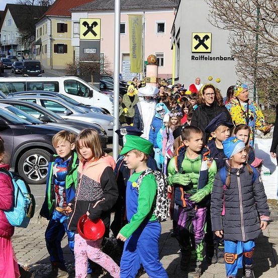 Eine Gruppe von Kindern in Kostümen geht mit geparkten Autos und einem Schild für Raiffeisenbank die Straße entlang. Sie werden von Erwachsenen begleitet und sind wahrscheinlich auf dem Weg zu einem Umzug oder einer Veranstaltung.