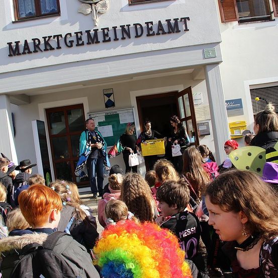 Eine Gruppe von Kindern, einige in Kostümen, versammelt sich vor dem Gebäude der Marktgemeindeamt. Ein Mann mit einem blauen Umhang steht am Eingang. Zwei Frauen im Gebäude tragen gelbe Kisten. Das Gebäude hat weiße Wände und Fenster.