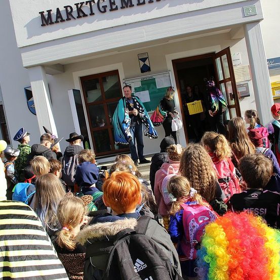 Eine Gruppe von Schülern versammelt sich vor einem Gebäude, 'MARKTGEMEIND'. Ein Mann steht am Eingang, und Menschen betreten das Gebäude. Einige Schüler tragen Kostüme, und Rucksäcke sind sichtbar.