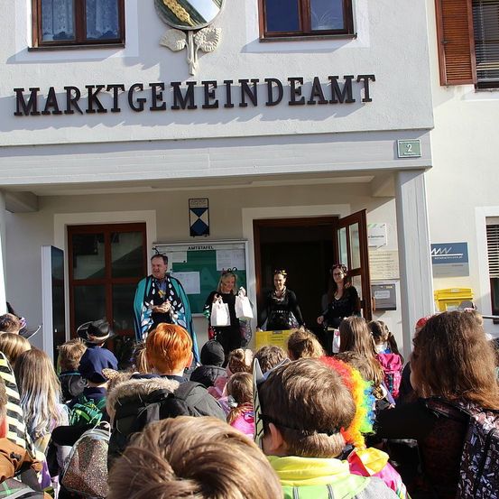 Eine Menschenmenge aus Kindern und Erwachsenen versammelt sich vor einem Gebäude mit der Aufschrift 'Marktgemeindeamt'. Zwei Personen mit Hüten stehen neben der offenen Tür.