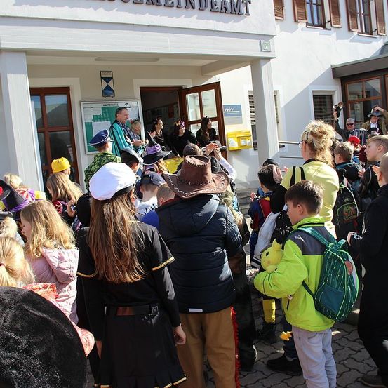Eine Gruppe von Kindern und Erwachsenen versammelt sich vor einem Gebäude, einige in Kostümen, einer hält ein Plüschtier. Leute machen Fotos und einige tragen Hüte.