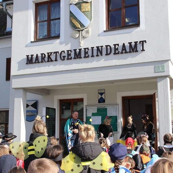 Eine Gruppe von Kindern in Bienenkostümen versammelt sich vor einem Gebäude. Der Mann in der Mitte hält ein Mikrofon. Das Schild am Gebäude lautet 'MARKTGEMEINDEAMT'. In der Nähe der Tür stehen zwei Frauen. Das Gebäude hat Fenster und ein schildartiges Emblem an der Wand.