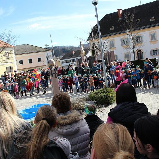 Eine Menschenmenge, einige in bunten Kostümen gekleidet, steht an einem sonnigen Tag auf einem Platz vor einem Gebäude. Eine Frau mit Pferdeschwanz und Brille schaut zu.