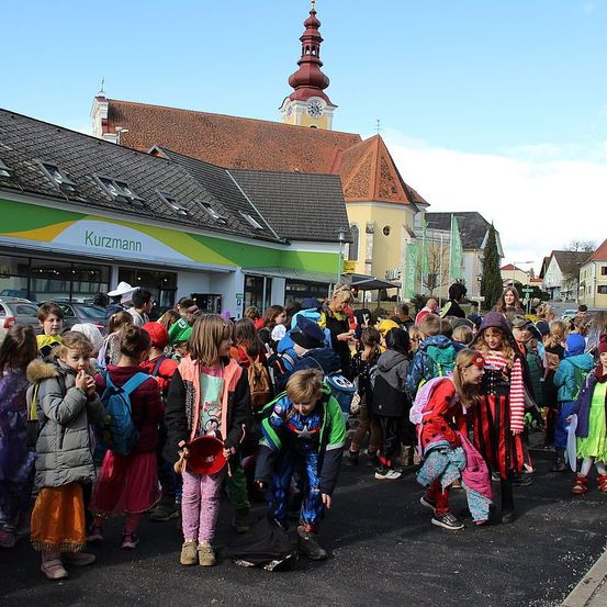 Eine Menschenmenge von Kindern in bunten Kostümen versammelt sich auf einem Stadtplatz, einige tragen Hüte und haben Rucksäcke dabei. Hinter ihnen sind ein Gebäude mit einem Glockenturm und eine Kirche mit einem roten Dach zu sehen.