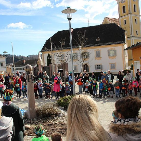 Eine Gruppe von Kindern und Erwachsenen versammelt sich vor einem Gebäude und trägt festliche Hüte. Die Szene spielt sich im Freien ab, mit einem klaren blauen Himmel.