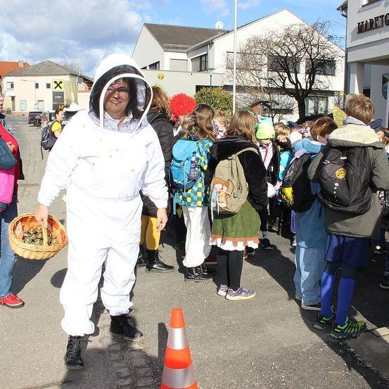Eine Gruppe von Menschen, einschließlich Kindern, steht im Freien. Ein Mann in einem Imkeranzug hält einen Korb. Hinter ihm tragen die Menschen Winterkleidung. Bäume und Gebäude sind im Hintergrund.