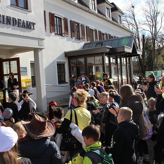 Eine Gruppe von Kindern in Kostümen versammelt sich vor einem Gebäude namens EinDeAmt. Sie klatschen und lächeln. Einige tragen Hüte, und eine Person in einem Bienenkostüm steht in der Nähe des Eingangs.