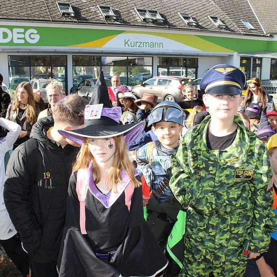 Kinder in Kostümen stehen vor einem Geschäft, 'Kurzmann', mit geparkten Autos davor. Eine Mädchen in einer Hexenmütze und ein Junge in einem Camouflage-Uniform stehen in der Mitte.