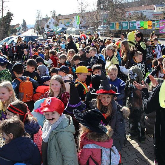 Eine große Menschenmenge von Schulkindern, einige in Kostümen, steht im Freien. Die Kinder stehen versammelt, einige tragen Hüte und Masken. Hinter ihnen sind Autos am Straßenrand geparkt.