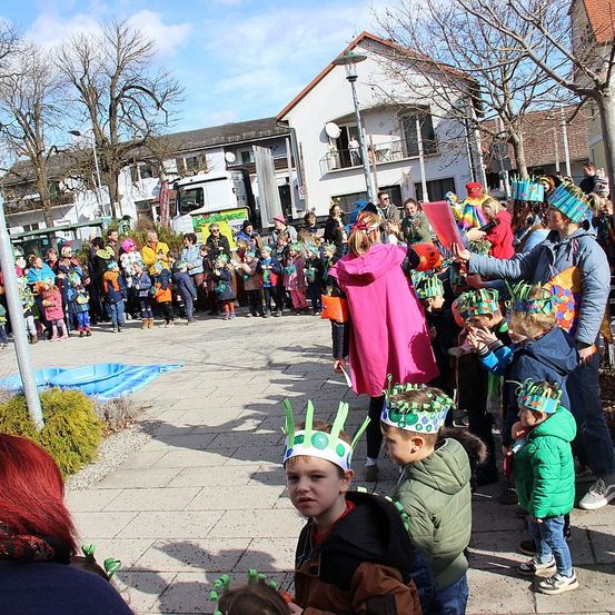 Eine Gruppe von Kindern und Erwachsenen versammelt sich im Freien in einer festlichen Umgebung und trägt bunte Hüte. Einige halten bunte Gegenstände in der Hand, andere schauen zu. Ein weißer Lastwagen ist in der Nähe geparkt.