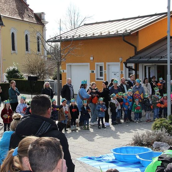 Eine Gruppe von Kindern und Erwachsenen versammelt sich vor einem gelben Gebäude mit einem Metalldach. Die Kinder tragen Blumenkränze. Zwei Erwachsene halten die Hände von Kindern, und blaue Becken liegen auf dem Boden.