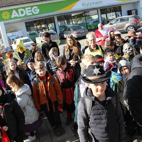 Eine Menschenmenge aus Kindern und Erwachsenen in verschiedenen Kostümen steht vor einem Geschäft mit dem Schild 'ADEG'. Einige tragen Superheldenkostüme, andere sind als Piraten gekleidet. Vor dem Geschäft sind Fahrzeuge geparkt.
