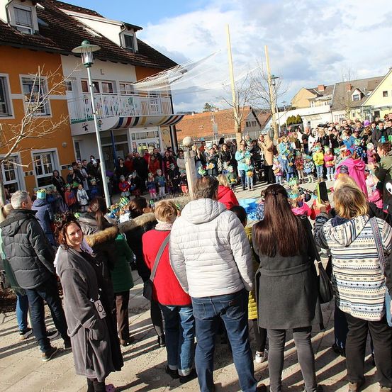 Eine Menschenmenge steht vor Gebäuden. Viele tragen Jacken und Mützen, einige haben Regenschirme. Sie beobachten eine Veranstaltung oder Aufführung. Bäume und Straßenlaternen sind in der Gegend.
