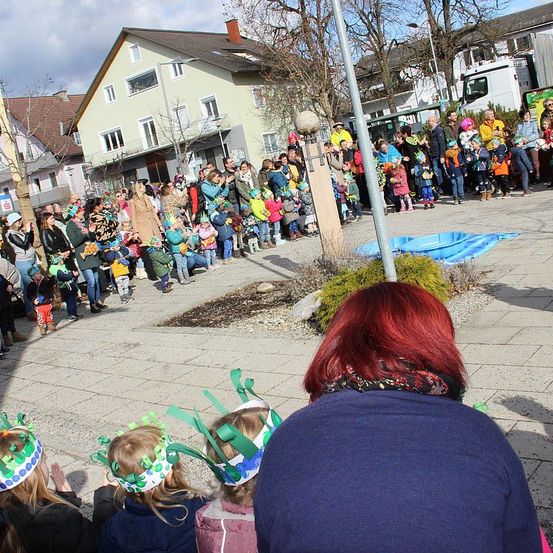Eine Menschenmenge, einschließlich Kinder und Erwachsene, versammelt sich auf einem Stadtplatz. Einige tragen festliche Hüte und einige halten Körbe. Eine Frau mit roten Haaren schaut sich die Menge an.