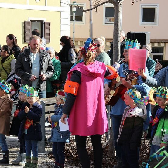 Eine Gruppe von Menschen, einschließlich Kindern, ist draußen versammelt. Einige Erwachsene stehen und beobachten, während Kinder Partyhüte tragen. Ein Mann mit einer Gitarre und Mikrofon spielt vor einem Baum. Gebäude mit Fenstern sind im Hintergrund.