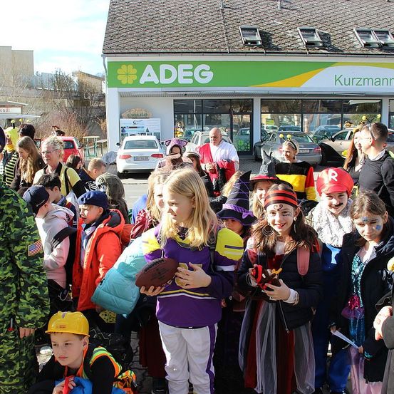 Eine Gruppe von Kindern in verschiedenen Kostümen, darunter ein Fußballspieler und eine Biene, versammelt sich vor einem Adeg-Laden, mit Erwachsenen und Autos im Hintergrund.