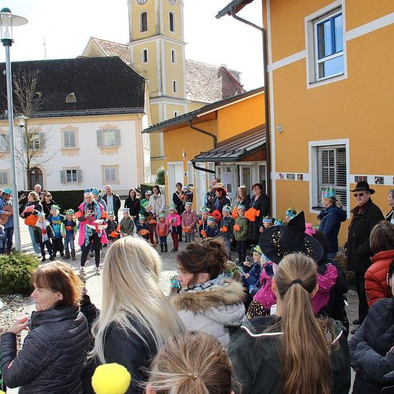 Eine Gruppe von Menschen in Kostümen steht vor einem Gebäude. Einige sind Kinder und andere Erwachsene. Eine Frau in der Front trägt einen Hut. Dahinter steht eine Straßenlampe und ein Gebäude mit einem Turm.